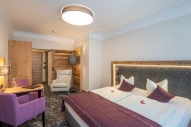 Das Bild zeigt ein Doppelzimmer im Westflügel mit neuer Zimmermöblierung, Boxspringbett, Tisch, 2 Sessel, Ohrensessel, Holz- und Lodenelemente