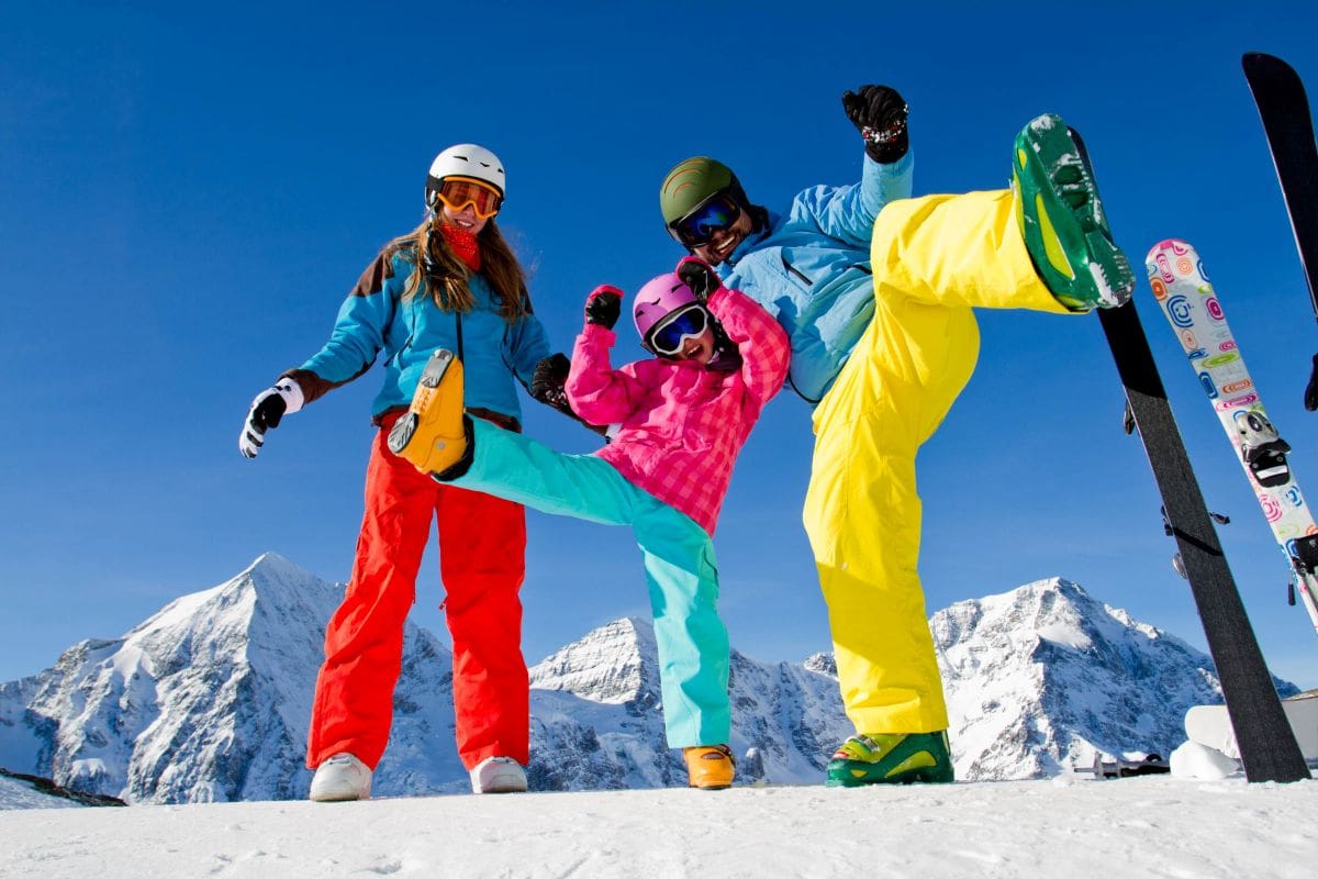 Eltern und Kind in farbenfroher Skikleidung stehen fröhlich mit Skiern vor einer verschneiten Bergkulisse