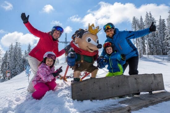 Family Spring Deals Ski Amadé - Familienskitag in Radstadt Familie mit zwei Kindern beim gemeinsamen Skitag in Radstadt neben dem Ski-Maskottchen Kinigl auf ener verschneiten Piste