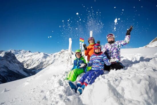 Family Spring Deals in Ski Amadé: Kinder fahren gratis Kinder in Skikleidung sitzen lachend im Schnee vor verschneiter Bergkulisse.