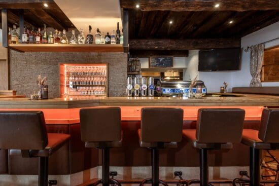 Gemütliche Bar im Restaurant Stegerbräu - Radstadt Barbereich im Restaurant-Stegerbräu in Radstadt mit beleuchtetem Gläserregal, Schank, Barhockern und historischen Holzbalken.